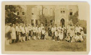 Photograph of a group of women on the lawn in front of a campus building. One woman is looking through a telescope.