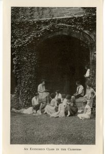 Photograph of students looking on, situated right near the ivy outside of the cloisters, as an instructor reads from an economics textbook.