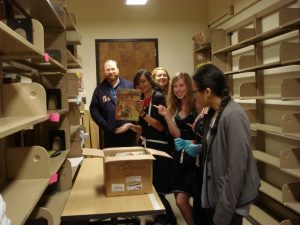 Catalogers and student employees, in processing room, hold up the last book above a shipping box.